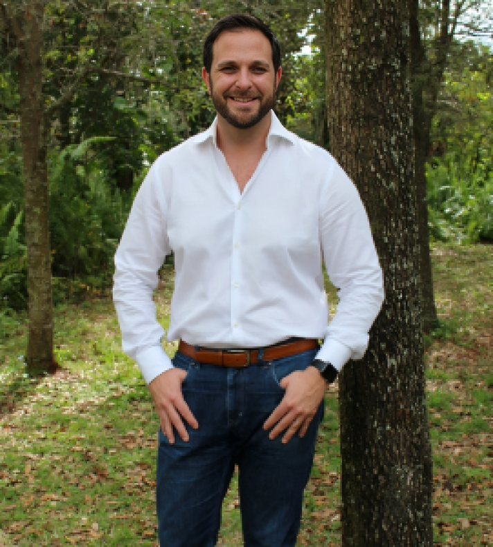 Professional man in white shirt and dark pants standing outdoors among trees.