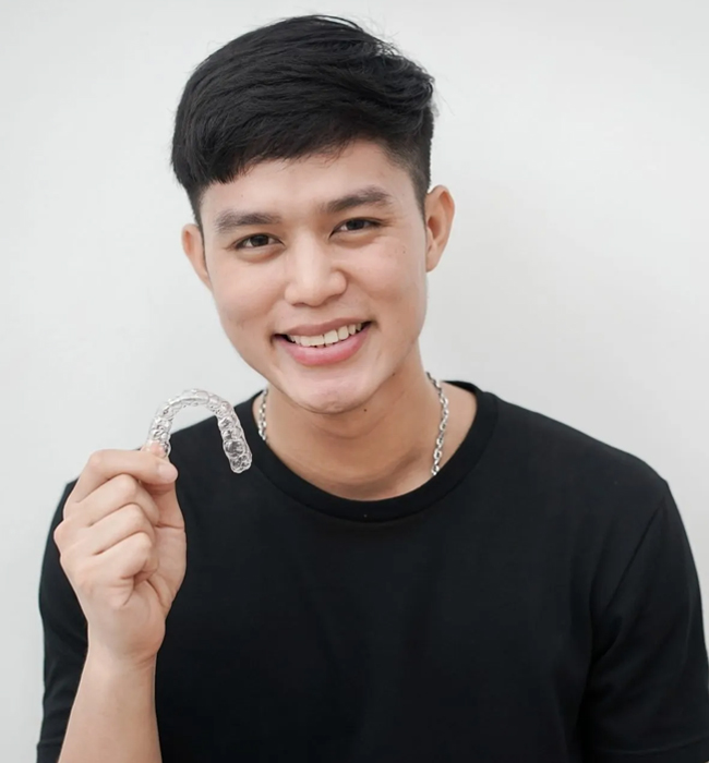 Young man smiling at camera, wearing black shirt, holding orthodontic braces, white background.