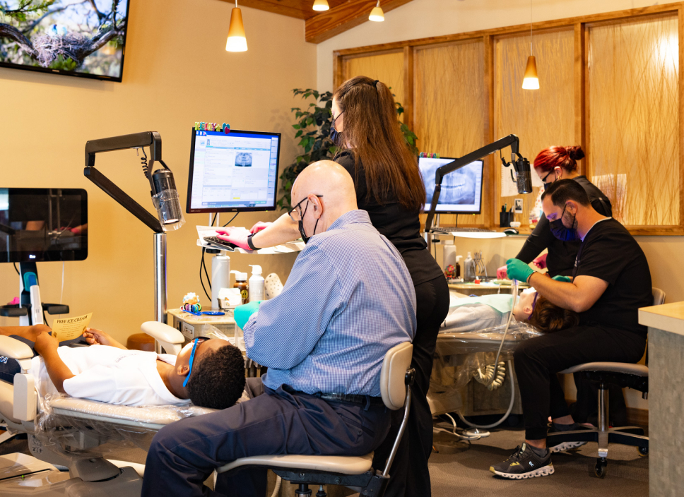 Orthodontist examines patient in modern dental office with staff members assisting at computer workstations.