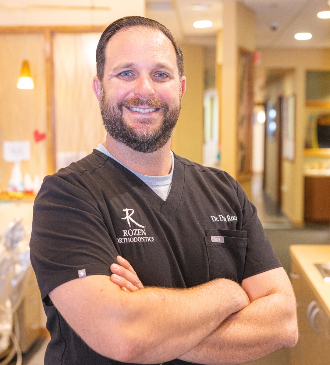 Smiling male orthodontist with arms crossed in modern dental office hallway wearing black scrubs.