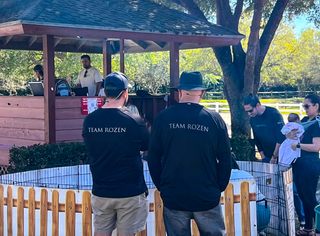Team Rozen members at outdoor event with booth and white picket fence, trees in background.