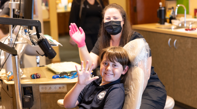 Child receiving orthodontic treatment in dental office while wearing mask, assisted by orthodontist.