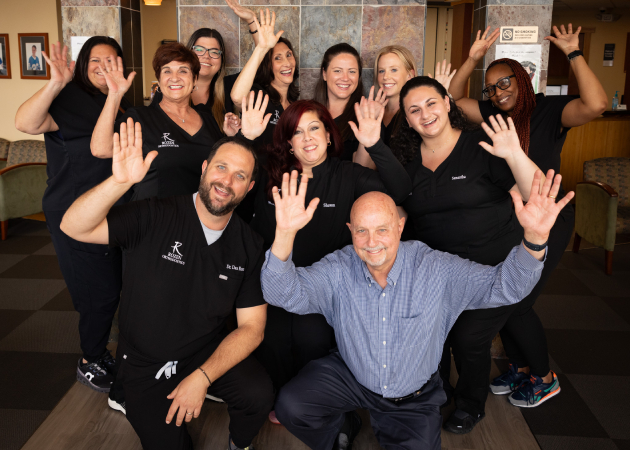 Rozen Orthodontics team members in black polos posing enthusiastically indoors with raised hands.