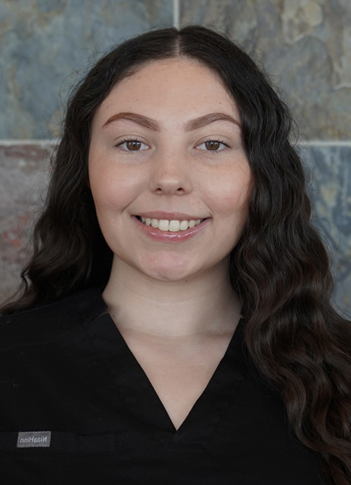 Professional headshot of smiling woman with long dark hair against gray tile background for orthodontics practice.