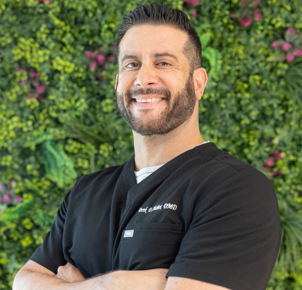 Smiling orthodontist in black scrubs poses against living plant wall background.