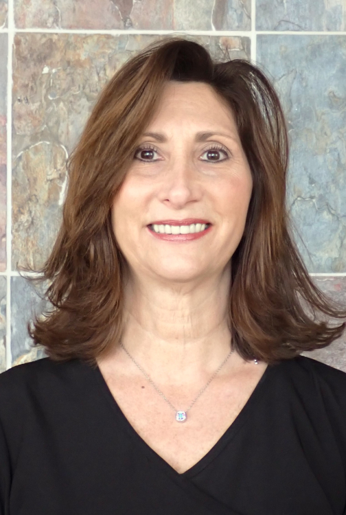 Professional headshot of a woman smiling in front of tiled background, wearing black top.