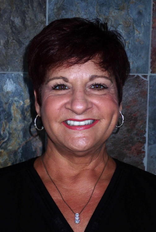 Professional headshot of smiling woman with short dark hair against blue tile background.