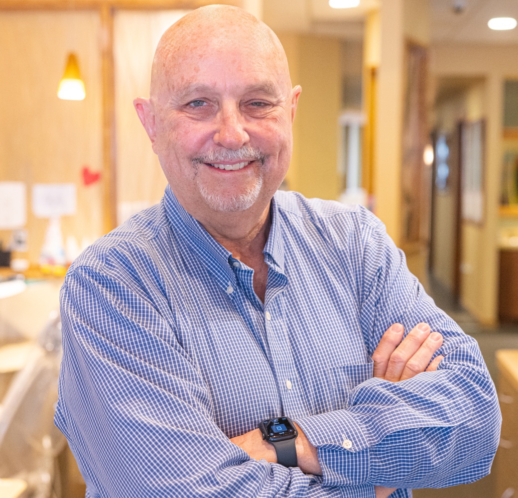 Smiling male orthodontist in blue shirt with crossed arms in modern dental office setting.