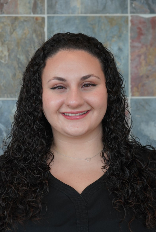 Professional headshot of a woman with long dark curly hair smiling at camera against tiled background.