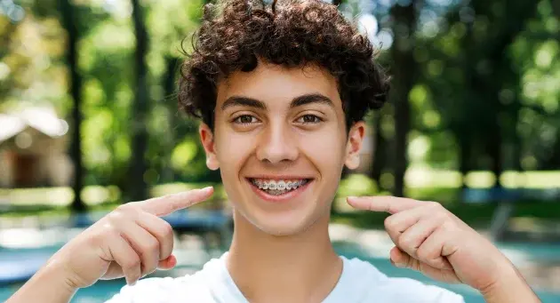 Smiling teenage boy with braces pointing to his teeth in a dental office setting.