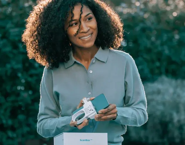 Smiling woman holding Scanbox dental monitoring kit against green foliage background.